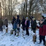 Groep mensen in winterkleding kijkt naar iets in een besneeuwd bos bij zonsondergang.
