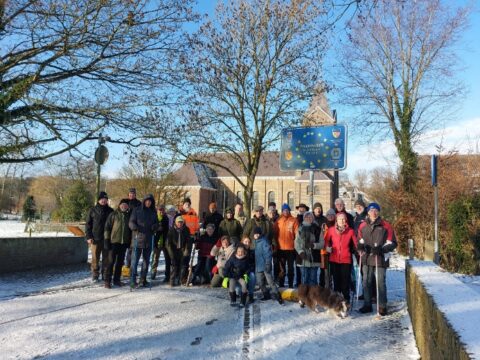 Groep mensen poseert buiten op een besneeuwde weg bij een kerk en een bord.
