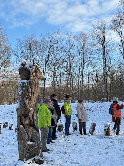 Groep mensen wandelt in besneeuwd bos, naast een houten sculptuur onder een blauwe lucht.