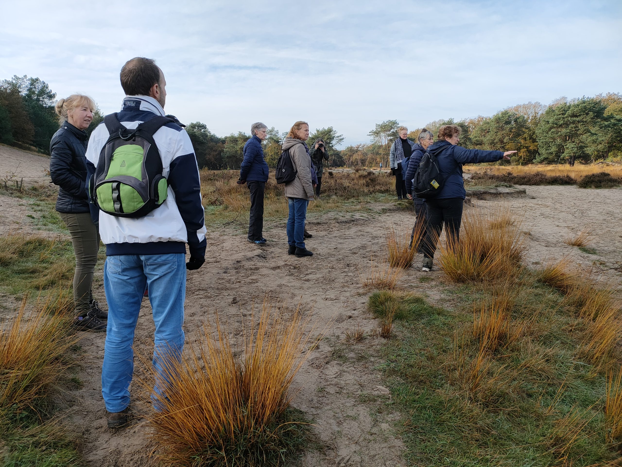 Groep mensen wandelt door een zandduinlandschap met gras en bomen in de verte.