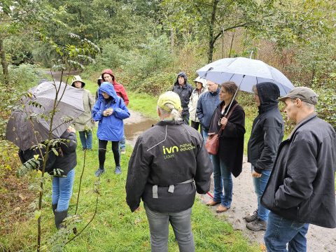Groep mensen met regenjassen en paraplu's tijdens een natuurwandeling in een bos.