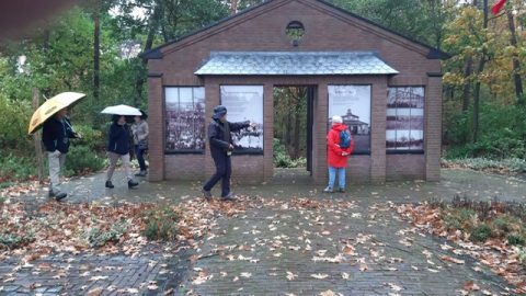 Groep mensen met paraplu's loopt langs een klein, bakstenen monument met zwart-wit foto's.