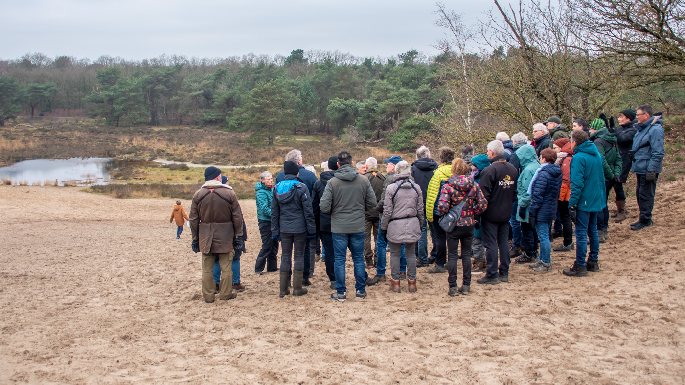 Groep mensen verzameld op een zanderige heuvel, met bos en een vijver op de achtergrond.