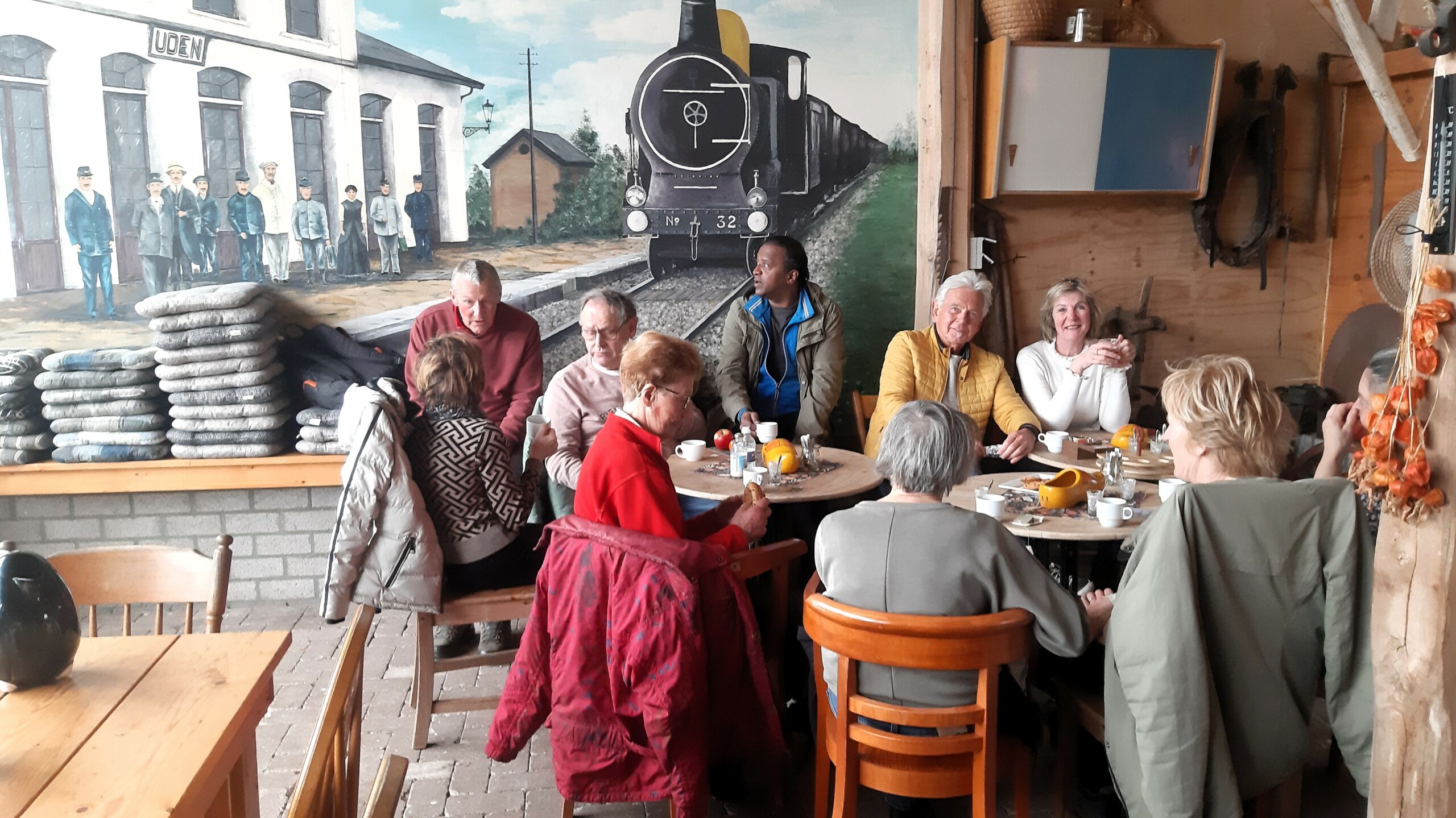 Groep mensen aan tafel met een muurschildering van een trein en station op de achtergrond.