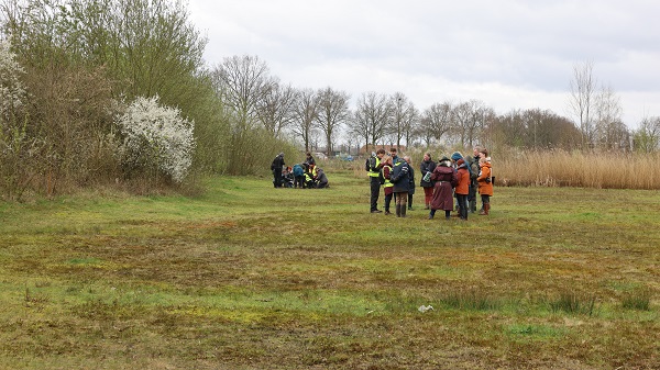 Groep mensen in een grasveld, omgeven door bomen en struiken, met een sombere lucht.