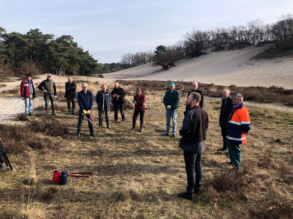 Groep mensen staat op een grasveld in een duinachtig landschap, enkele dragen handschoenen en gereedschap.