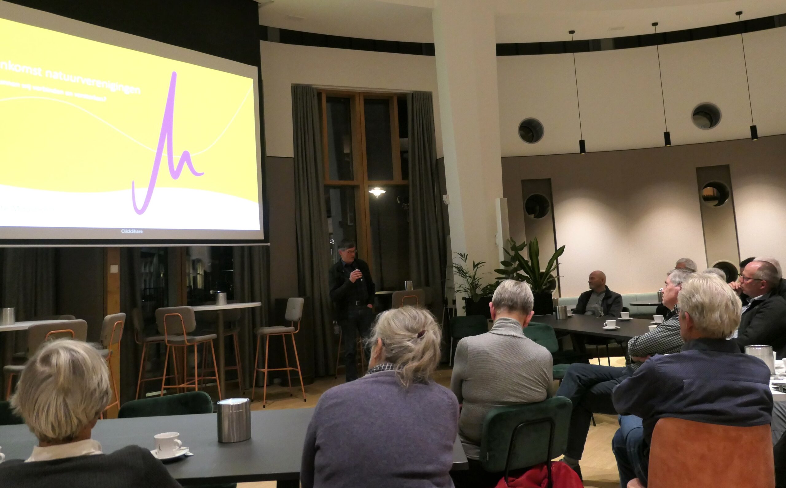 Een presentatie in een conferentieruimte met publiek en spreker. Scherm toont gele dia.