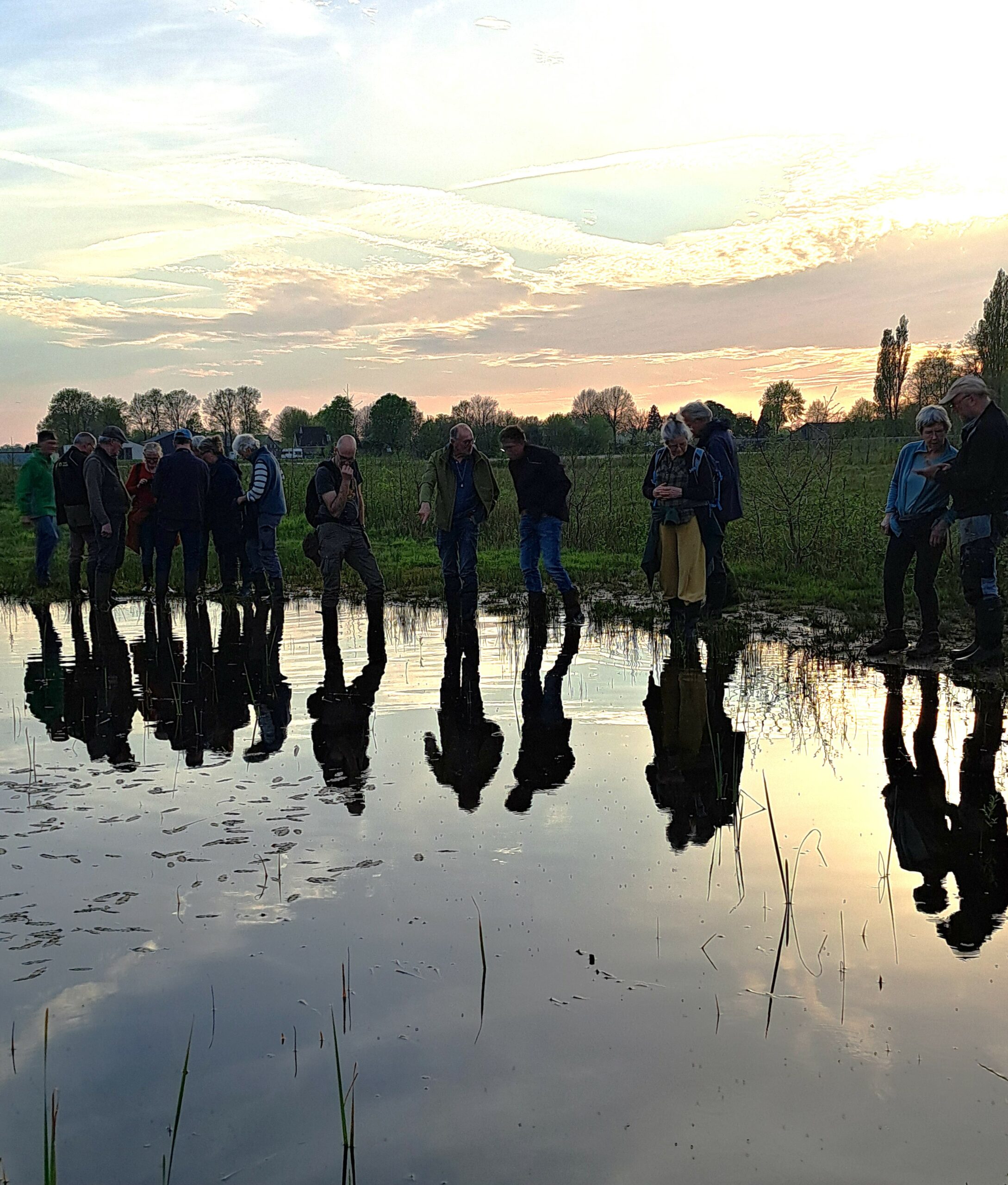 Groep mensen staat bij een vijver tijdens zonsondergang, reflecties zichtbaar in het water.