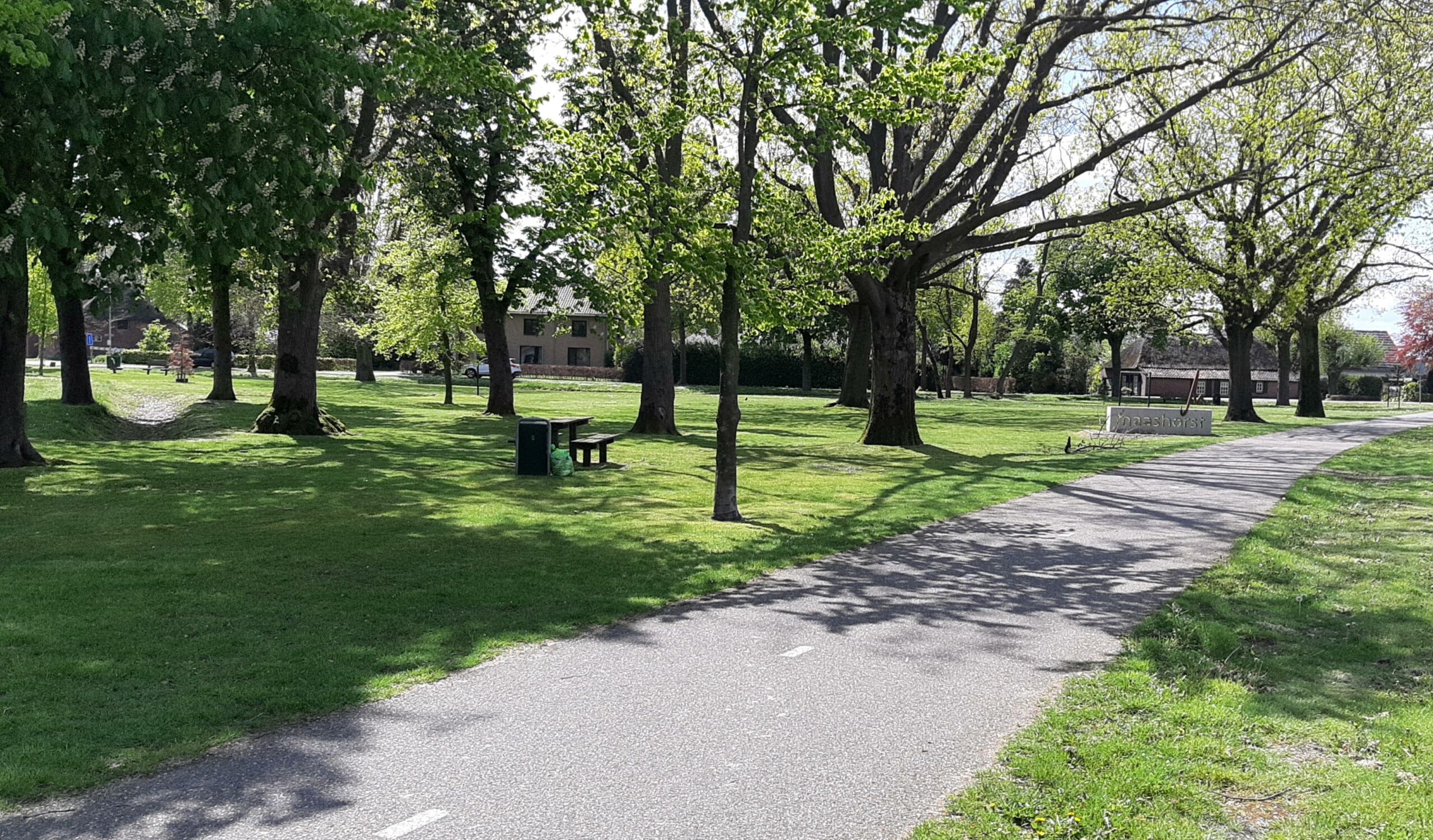 Park met grasveld, bomen, bankjes en een fietspad op een zonnige dag; huis en bord op de achtergrond.