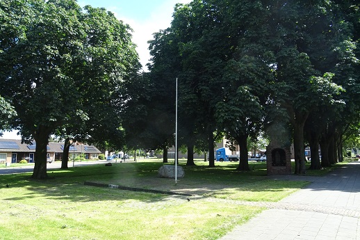 Groen park met bomen, een lege vlaggenmast en een verhard pad rechts.