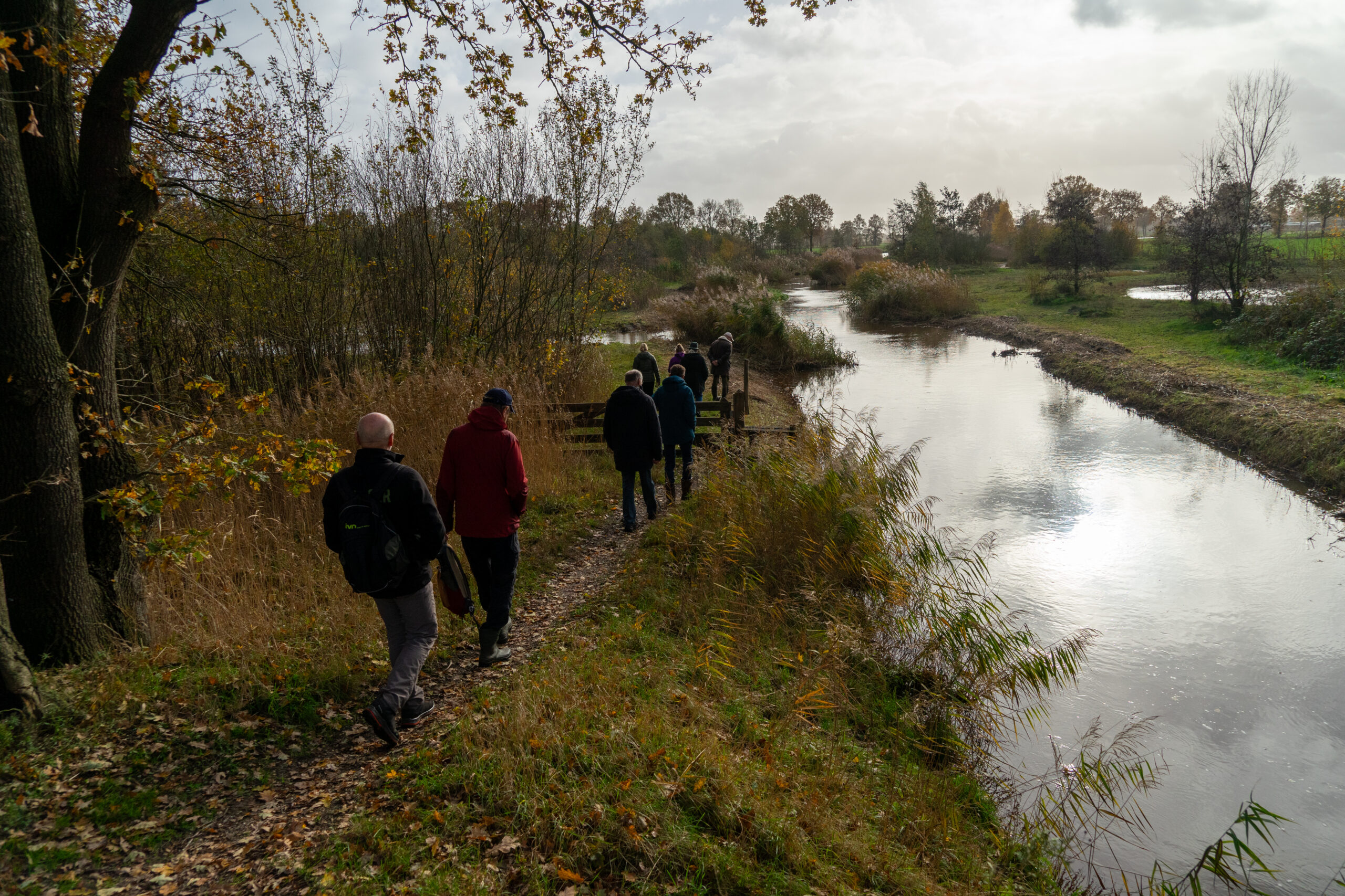 Mensen wandelen langs een rivieroever te midden van herfstbomen en grasland.
