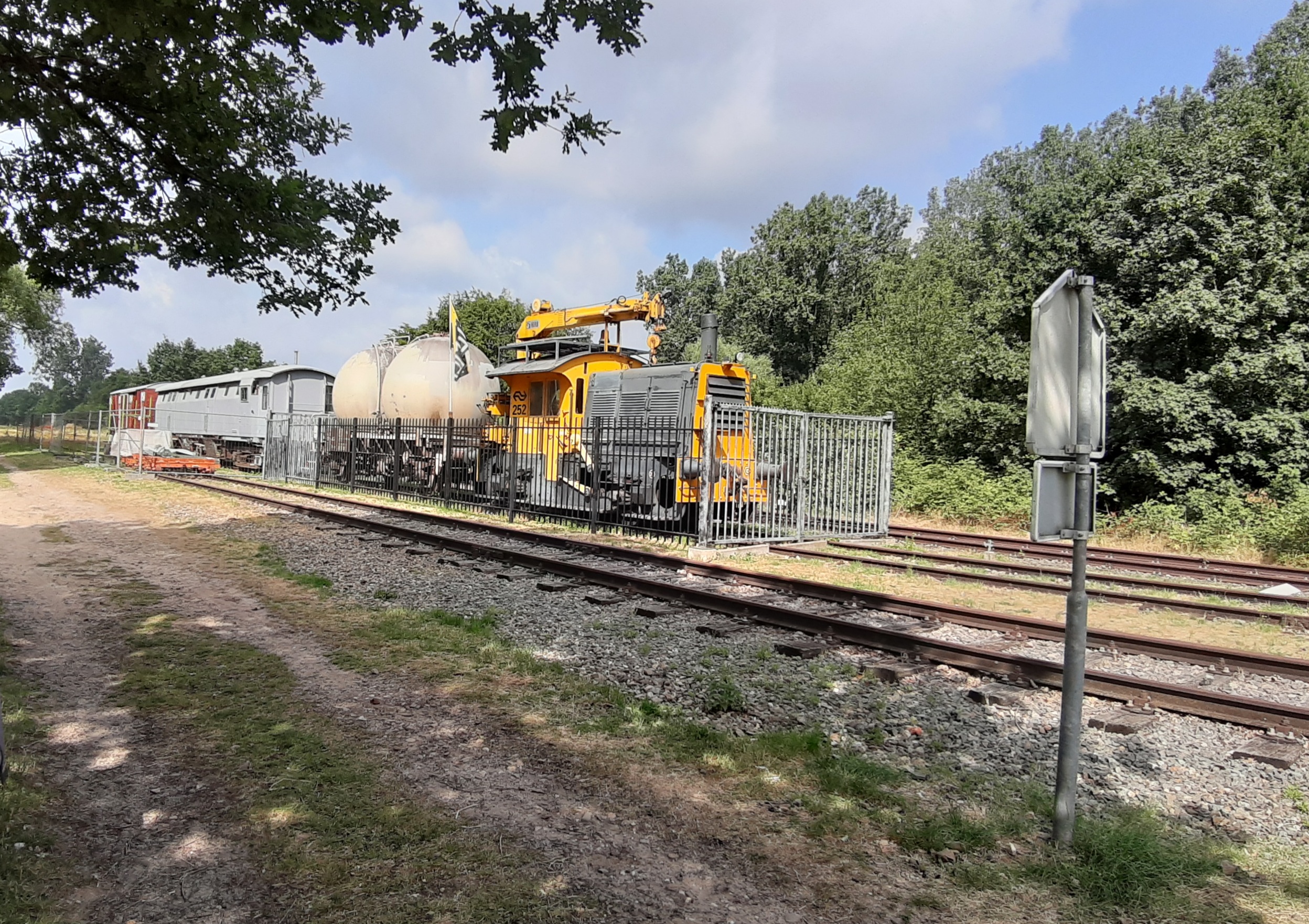 Een gele onderhoudslocomotief naast een spoor, omringd door bomen en een hek.