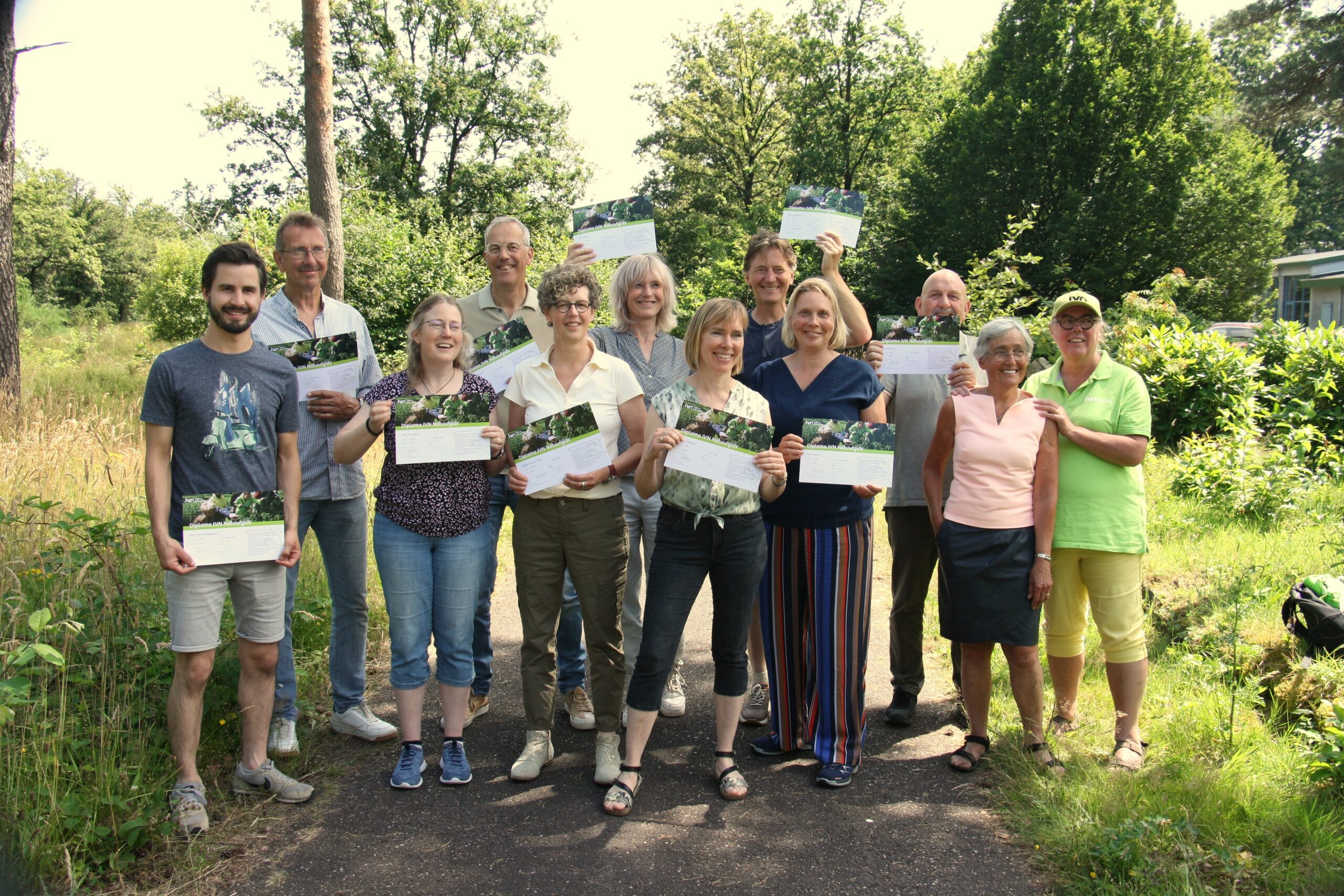 Groep van elf mensen buiten, glimlachend en met certificaten in handen, omgeven door groene natuur.