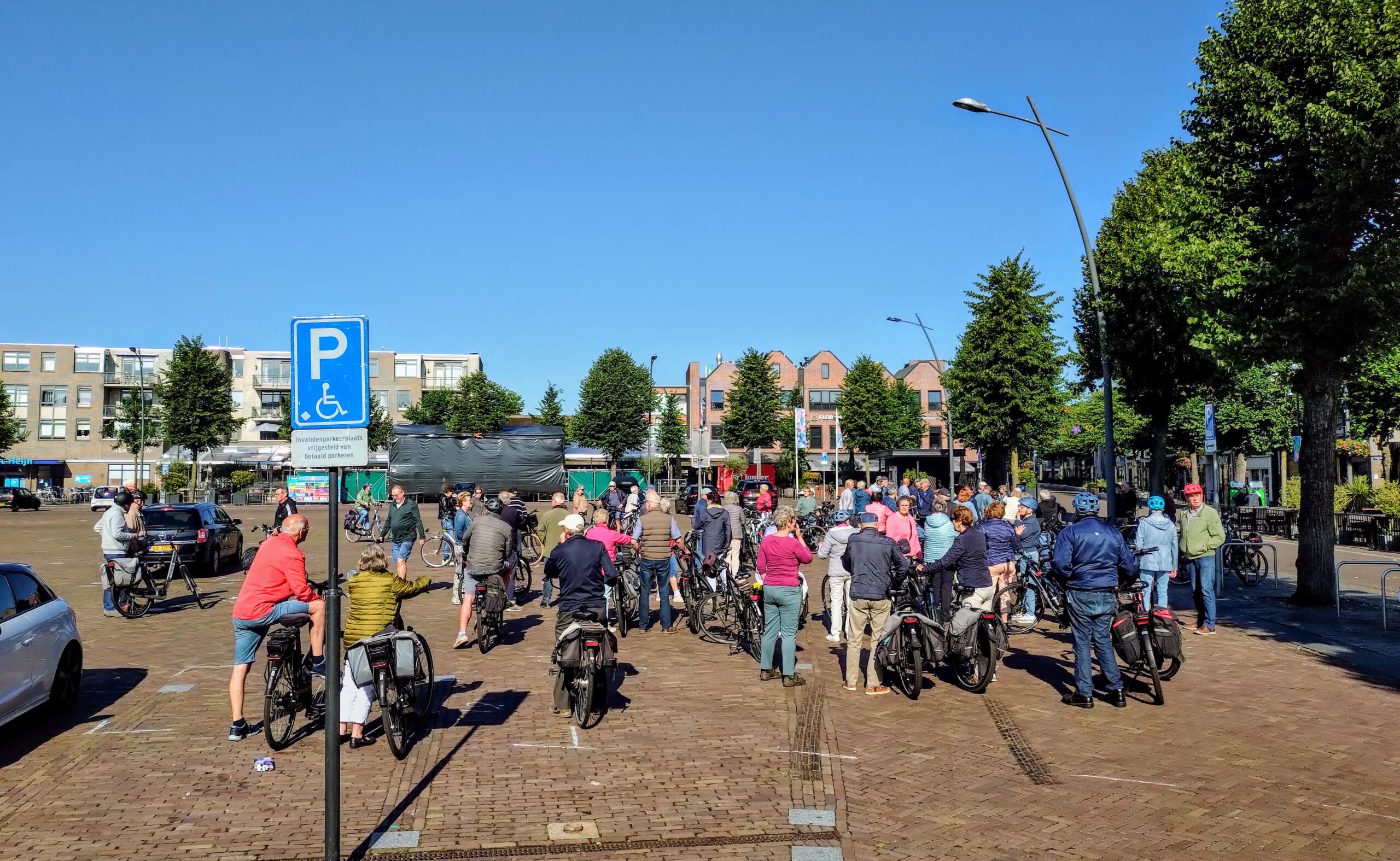 Groep mensen op fietsen verzamelt zich op een plein bij een parkeerbord voor gehandicapten.