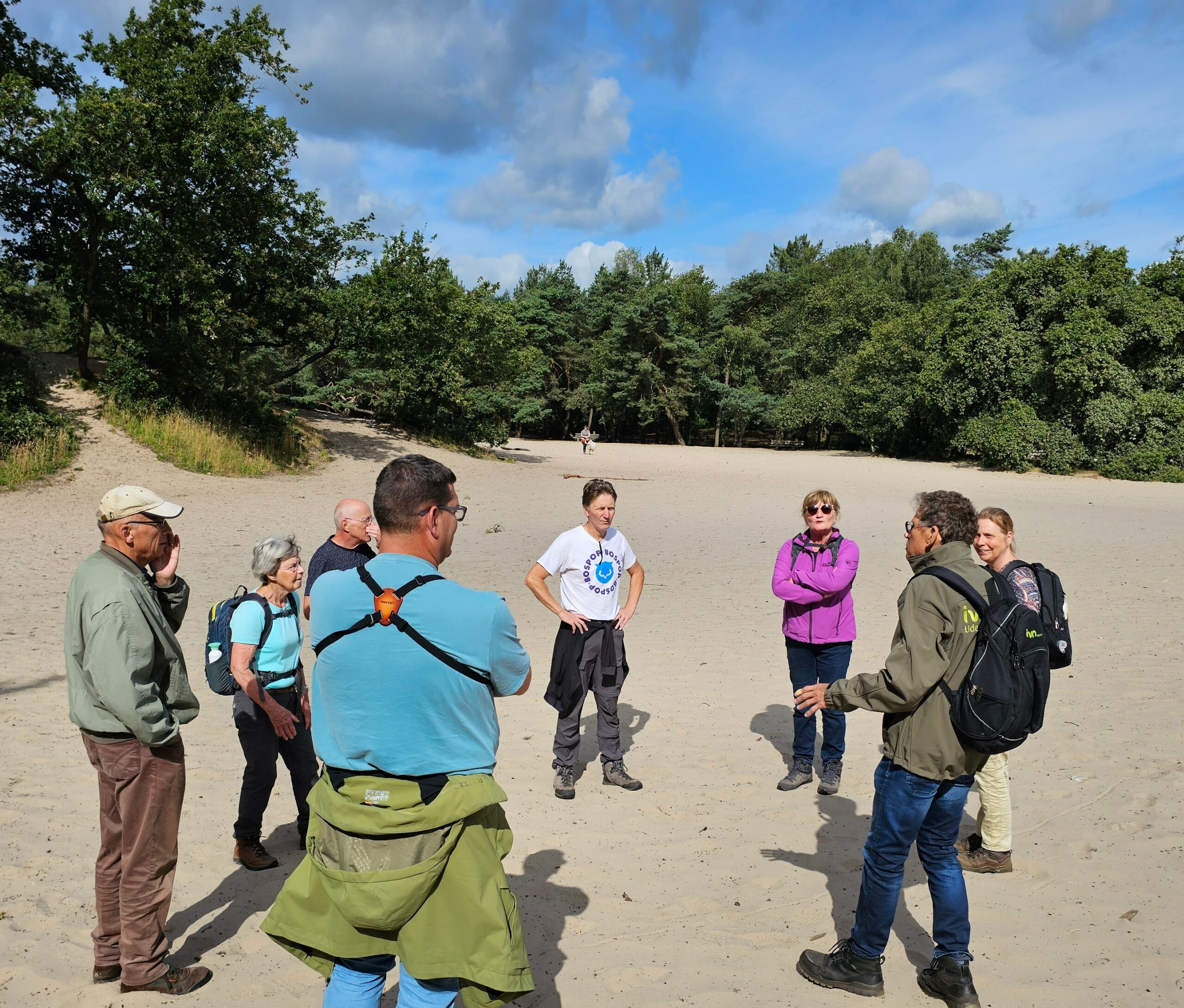 Groep mensen staat op een zandpad in een bosgebied tijdens een zonnige dag, in gesprek met elkaar.