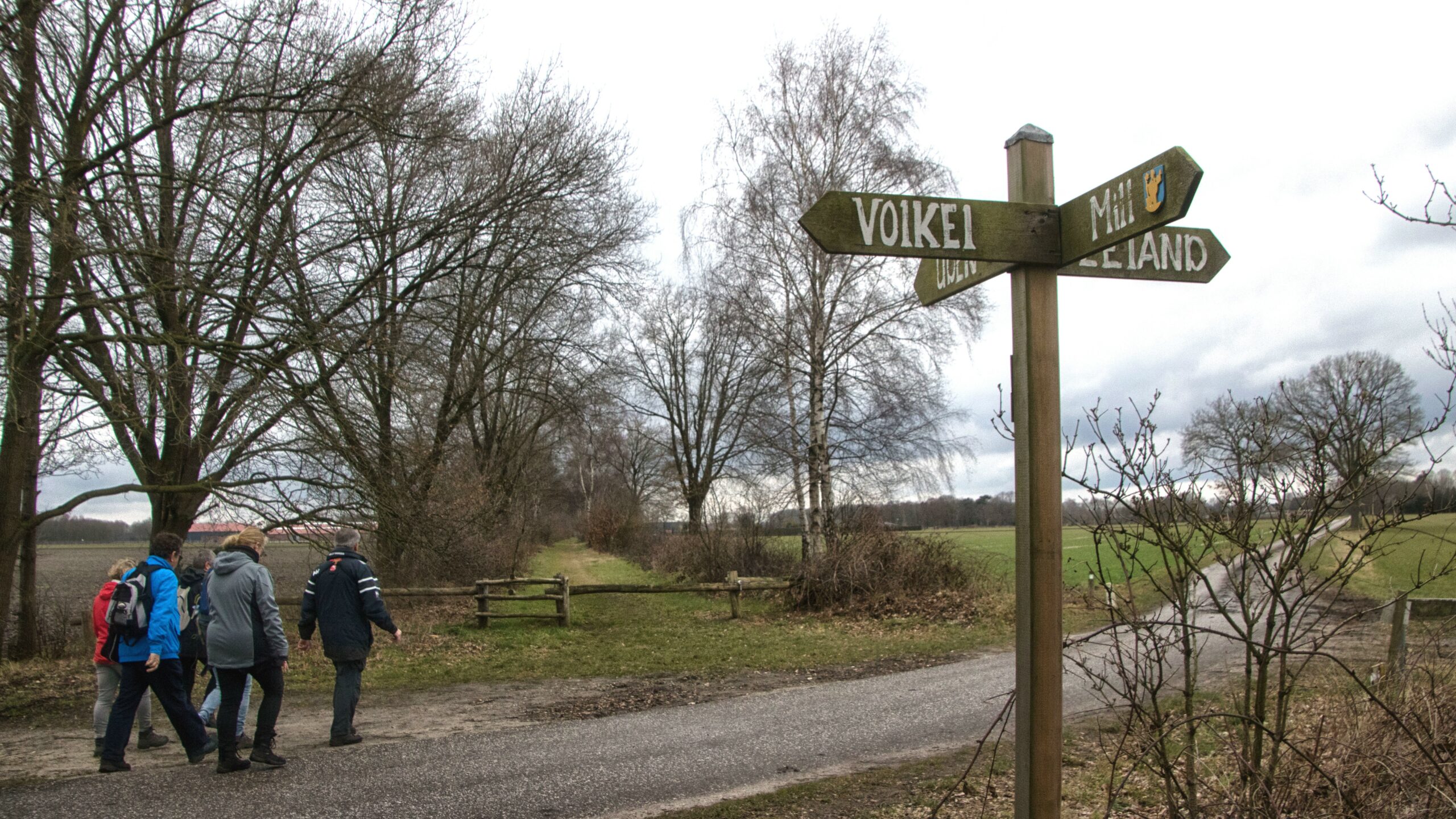 Wandelaars passeren een houten bord met richtingaanwijzers, omgeven door kale bomen en velden.