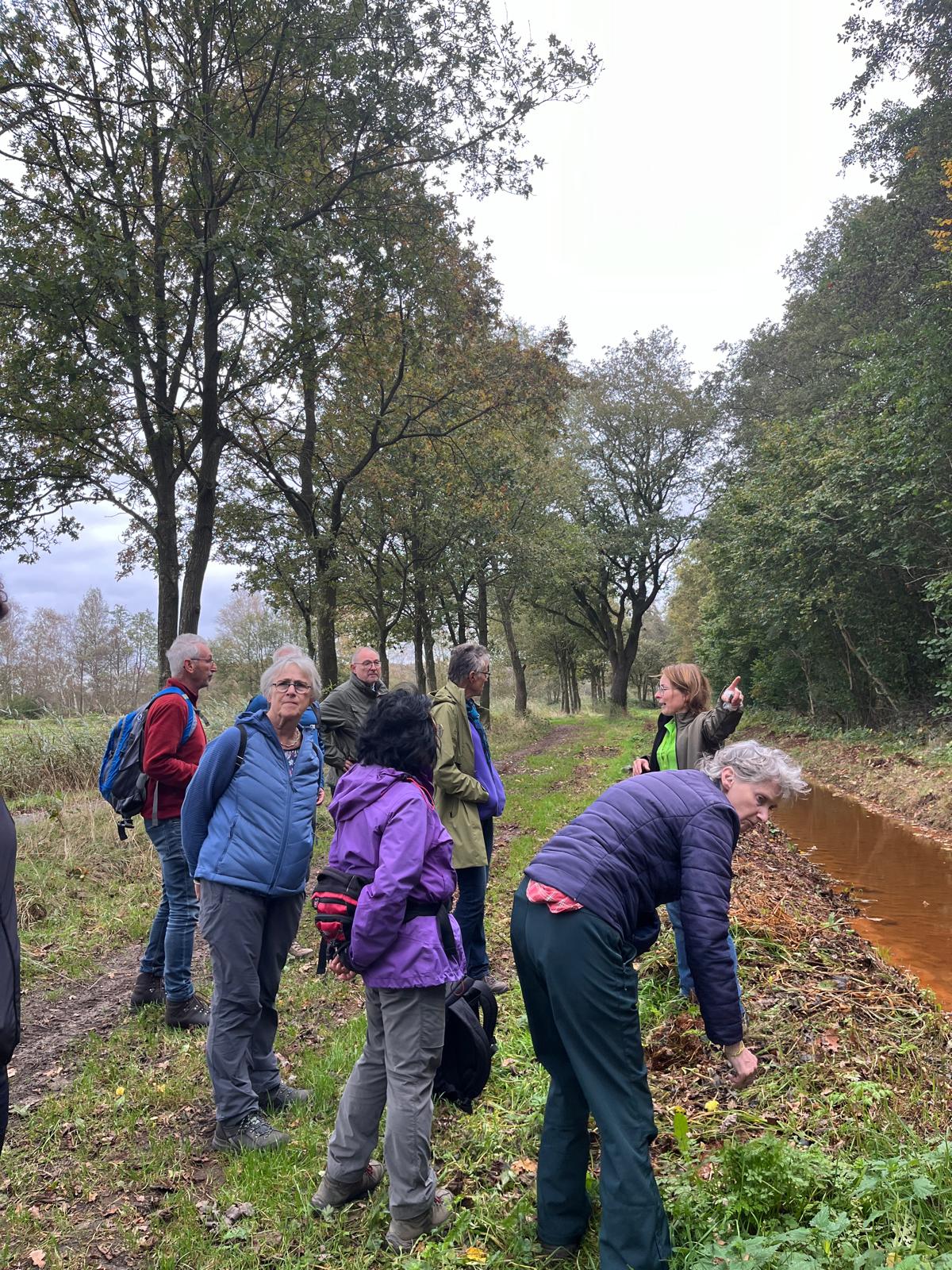 Groep mensen wandelt langs een boomrijk pad met een smalle sloot aan de rechterkant.