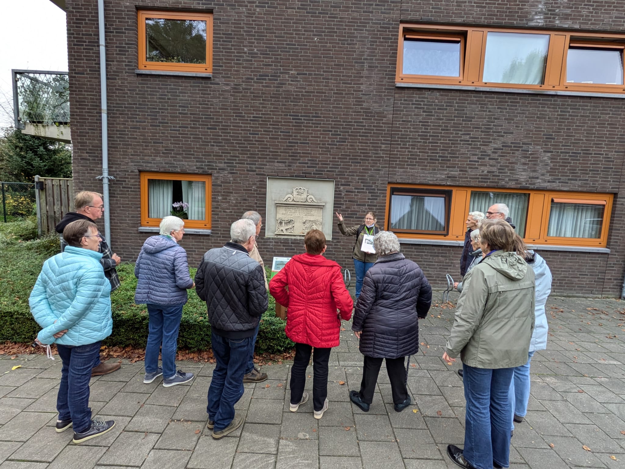 Een groep mensen luistert naar een gids bij een gevelsteen aan een bakstenen muur.