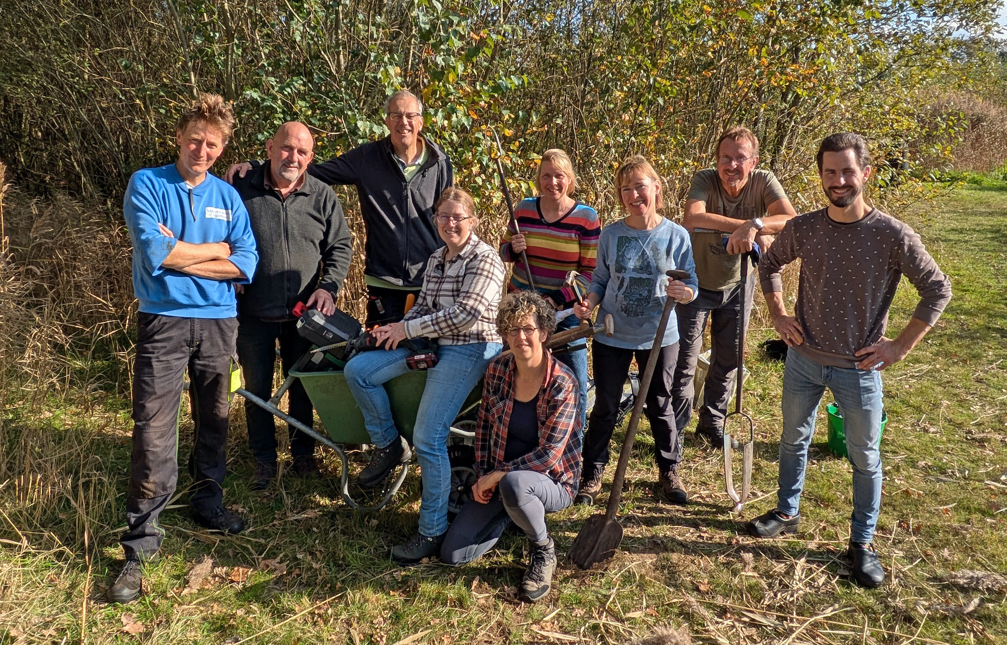 Groep mensen op een zonnige dag in de natuur met tuingereedschap en een kruiwagen.