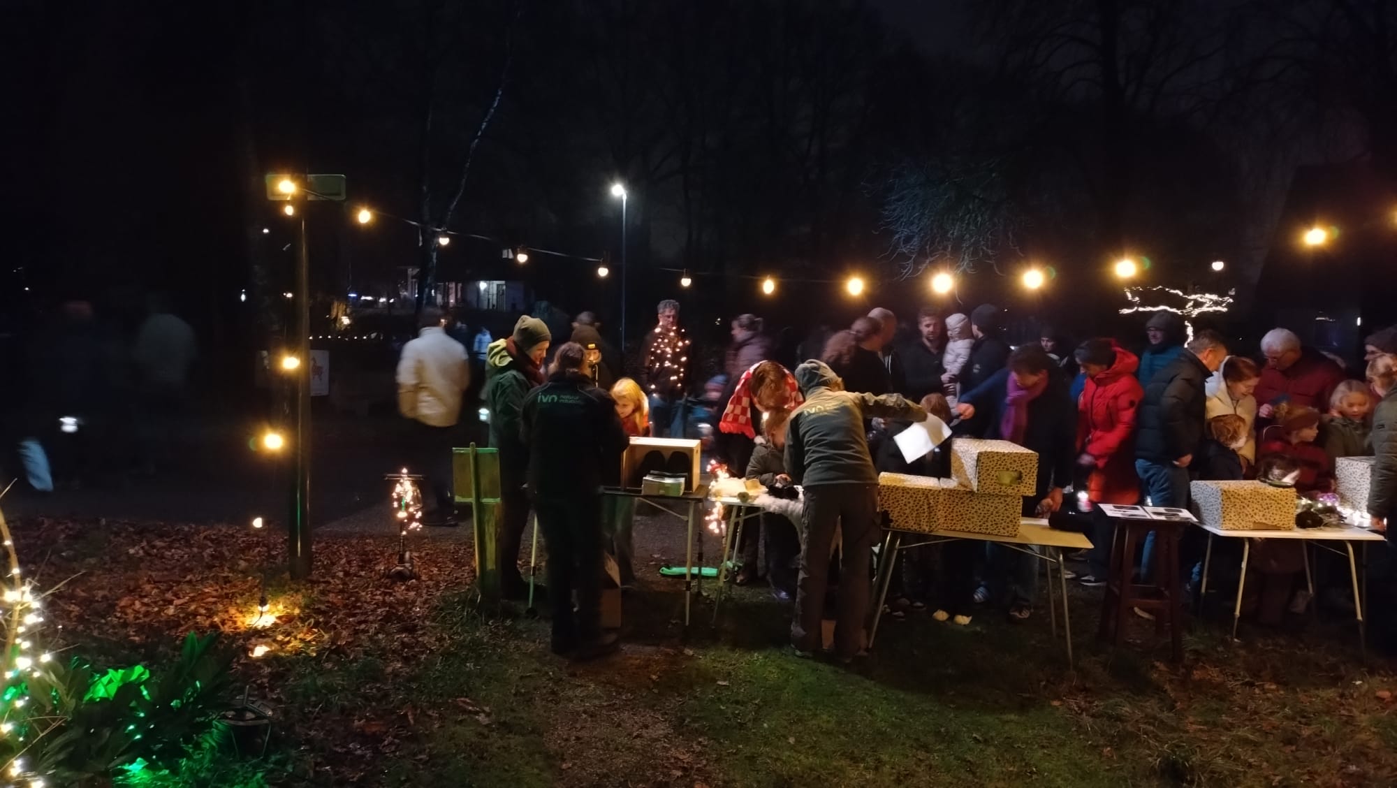 Groep mensen bij een avondmarkt met kerstverlichting, tafels en cadeaus.