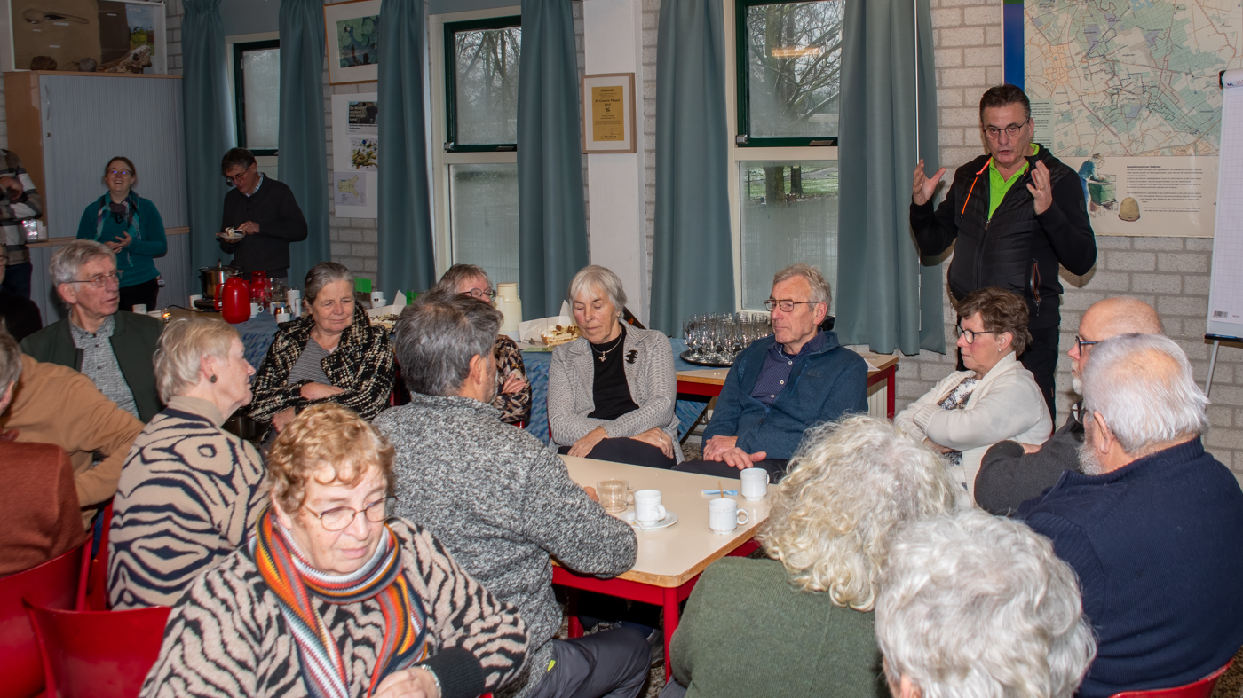 Groep mensen luistert naar een spreker in een zaal met kaarten en gordijnen. Koffiekopjes op tafel.