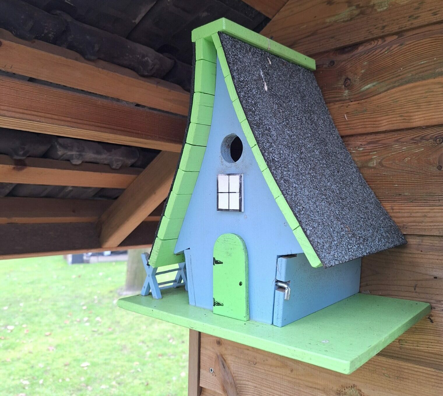 Een blauwe en groene sprookjesachtige vogelhuisje met schuin dak hangt aan een houten wand.
