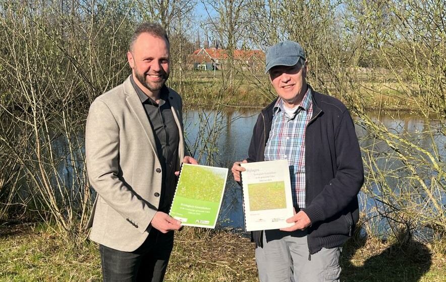 Twee mannen houden rapporten vast bij een vijver, met bomen en een huis op de achtergrond.