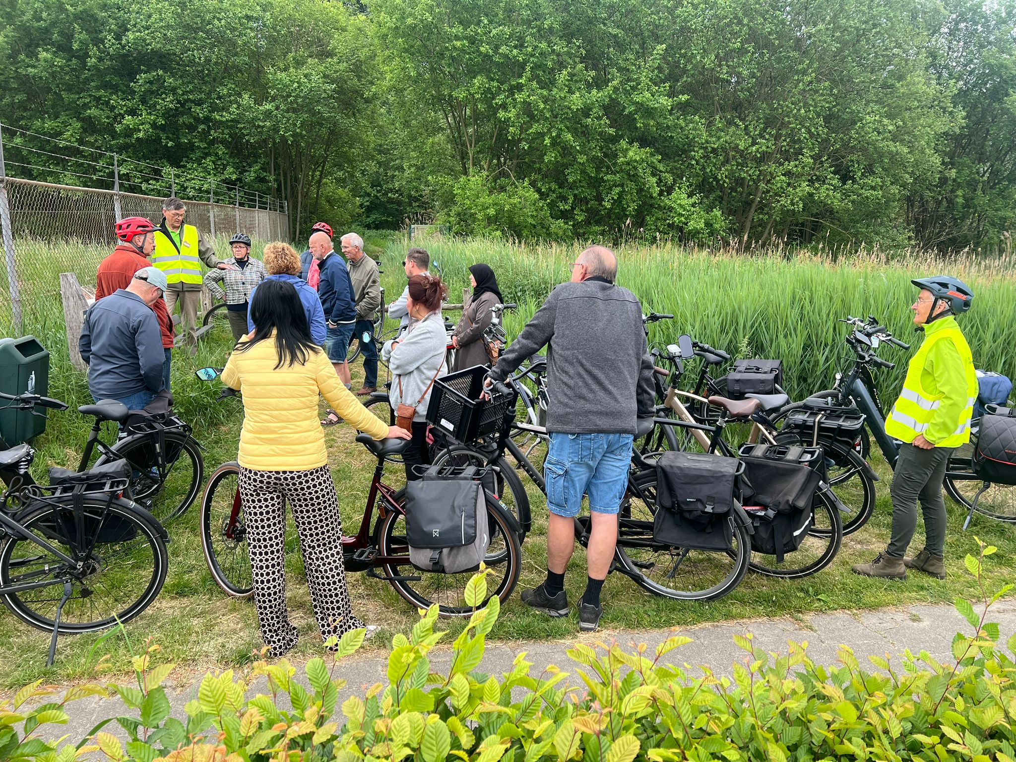 Groep fietsers verzameld, luisterend naar gids in gele veiligheidsvest, naast groen landschap.