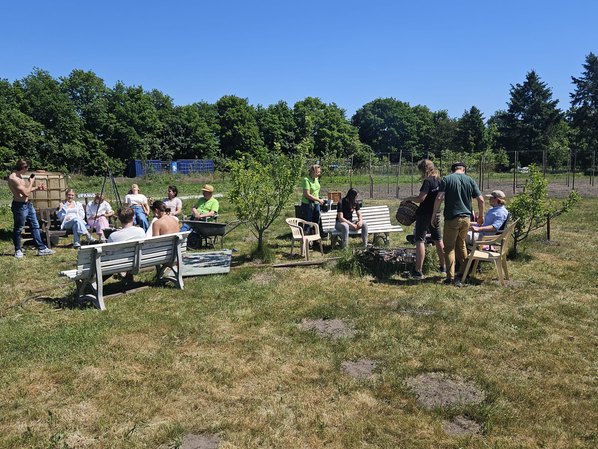 Groep mensen zit en staat bij banken in een tuin, omgeven door gras en bomen, op een zonnige dag.