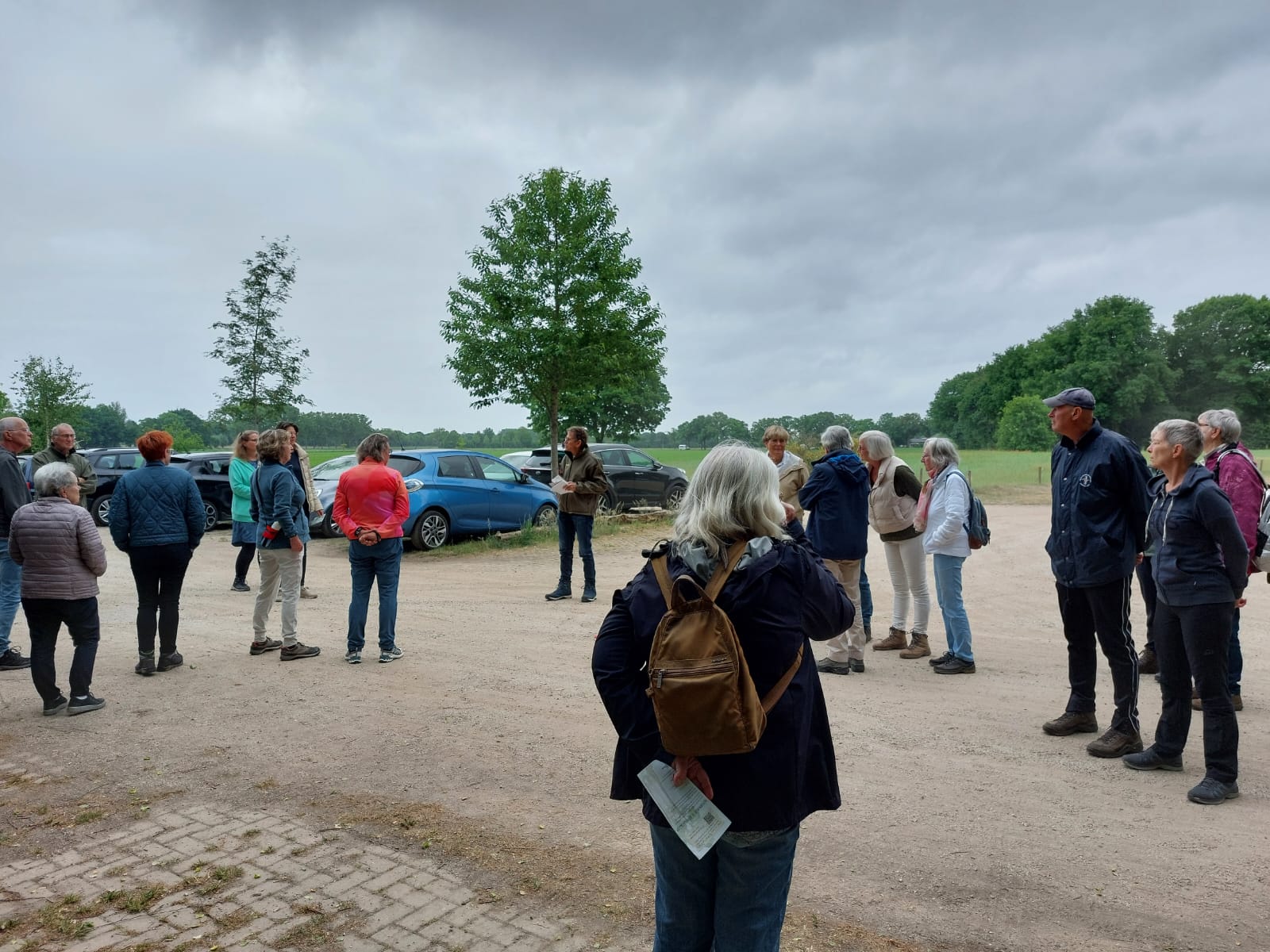 Groep mensen luistert naar een spreker buiten op een parkeerplaats met auto's en bomen. Mensen dragen jassen.