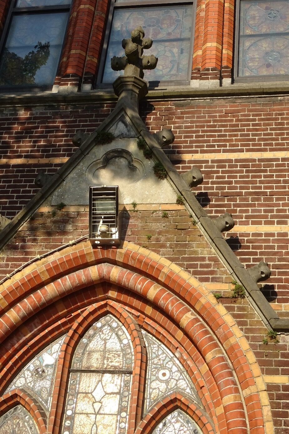 Brikstenen gevel met glas-in-loodramen en decoratieve kruisvormige sculptuur.