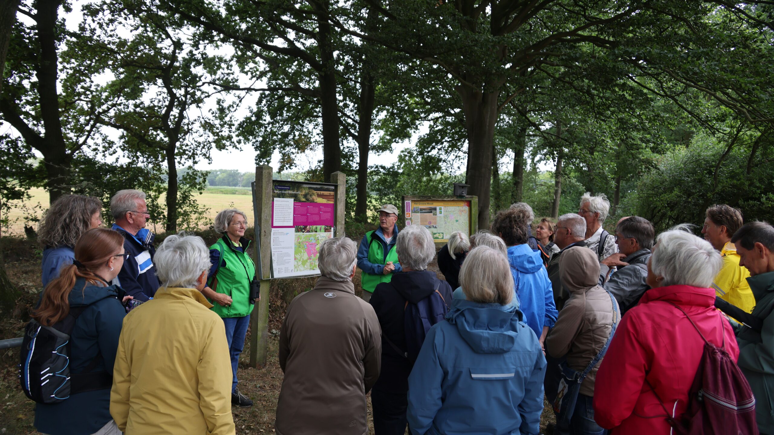 Groep mensen luistert naar gids bij informatieborden in bosrijke omgeving.