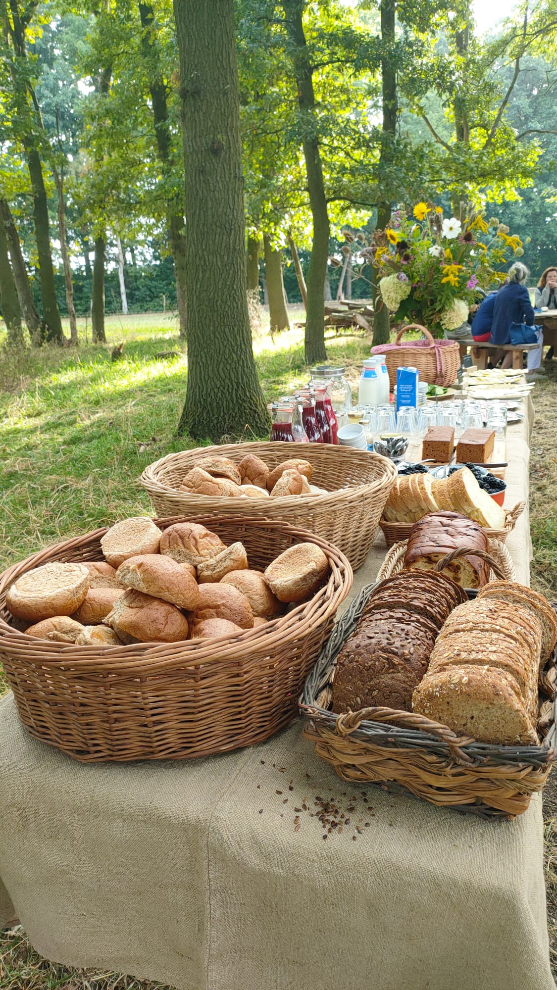Picknicktafel in bos met broodmanden, gebak, bloemen en mensen op de achtergrond.