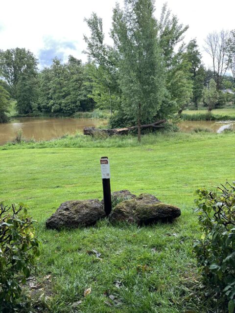 Groen grasveld, boomstammen bij een rustige vijver. Bordje in het midden bij grote stenen.