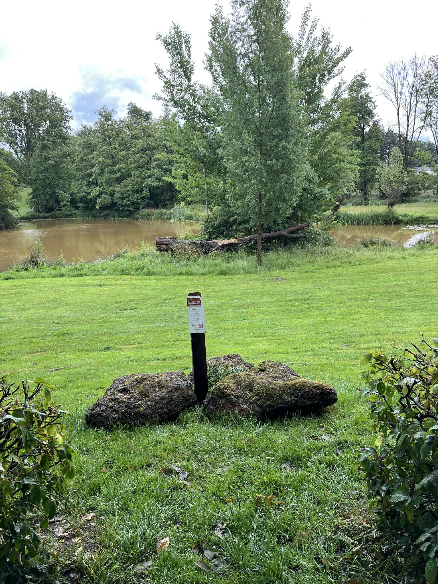 Groen grasveld, boomstammen bij een rustige vijver. Bordje in het midden bij grote stenen.