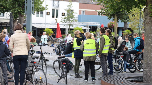 Groep mensen met fietsen, sommigen dragen gele hesjes, bijeen op een plein met bomen en gebouwen.