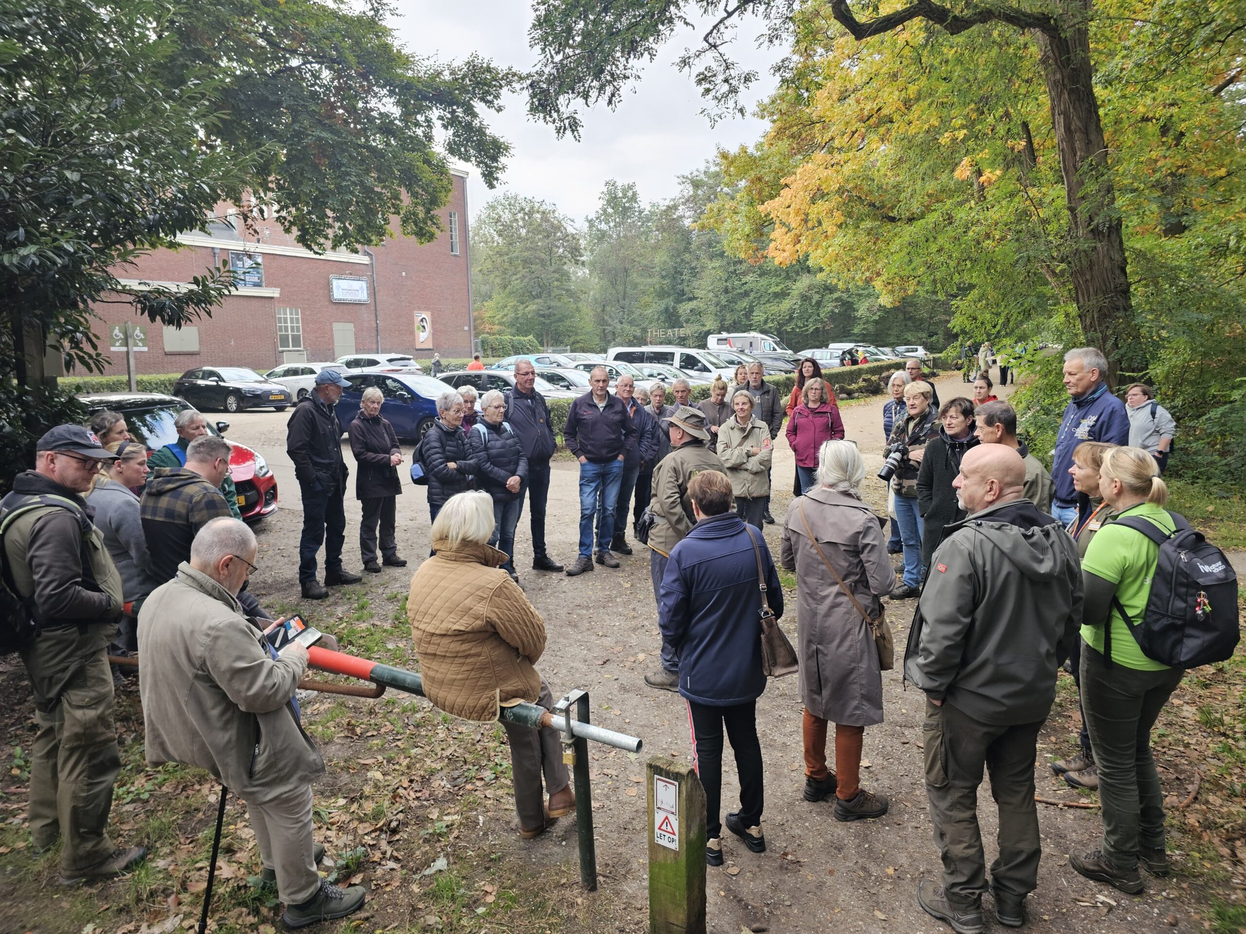 Groep mensen verzameld in een bosrijke omgeving bij auto's en een gebouw.