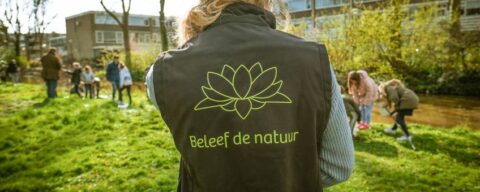 Persoon in natuurgebied met logo "Beleef de natuur", kinderen op achtergrond spelen buiten.