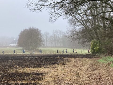Wandelaars lopen door een mistig landschap met bomen en een huis op de achtergrond.