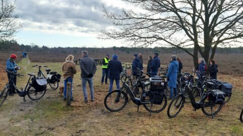 Groep mensen met fietsen in een natuurgebied, luisterend naar een spreker in een geel hesje.