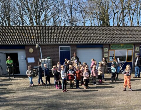 Groep kinderen en volwassenen voor een gebouw met opschrift "In Hof van de Toekomst" op een zonnige dag.