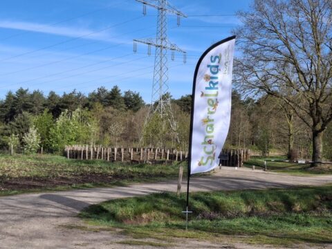Vlag met "Schatrelkids" bij een bospad, met bomen en elektriciteitsmast op de achtergrond.