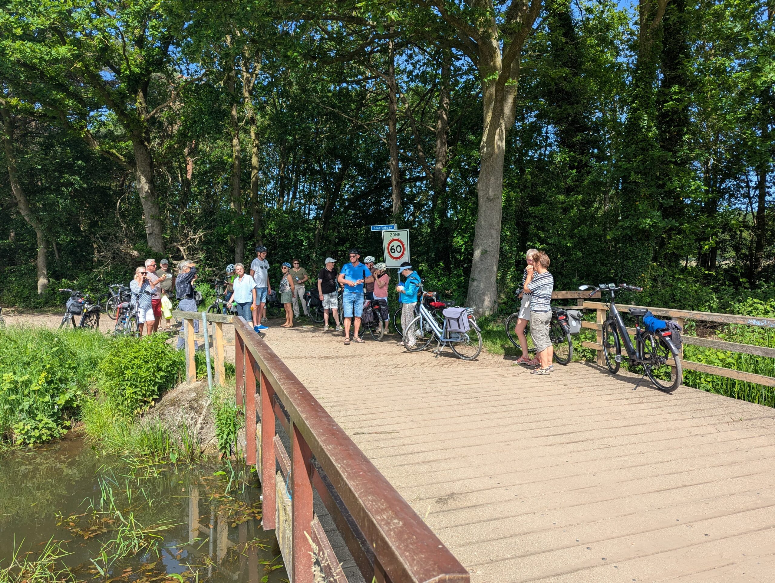 Groep fietsers op een brug in een bosrijke omgeving, met fietsen geparkeerd en een snelheidsbord.