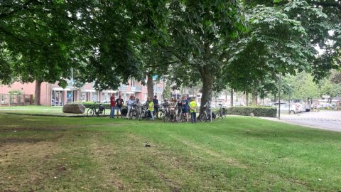 Groep mensen met fietsen onder bomen in een park, naast een parkeerplaats.