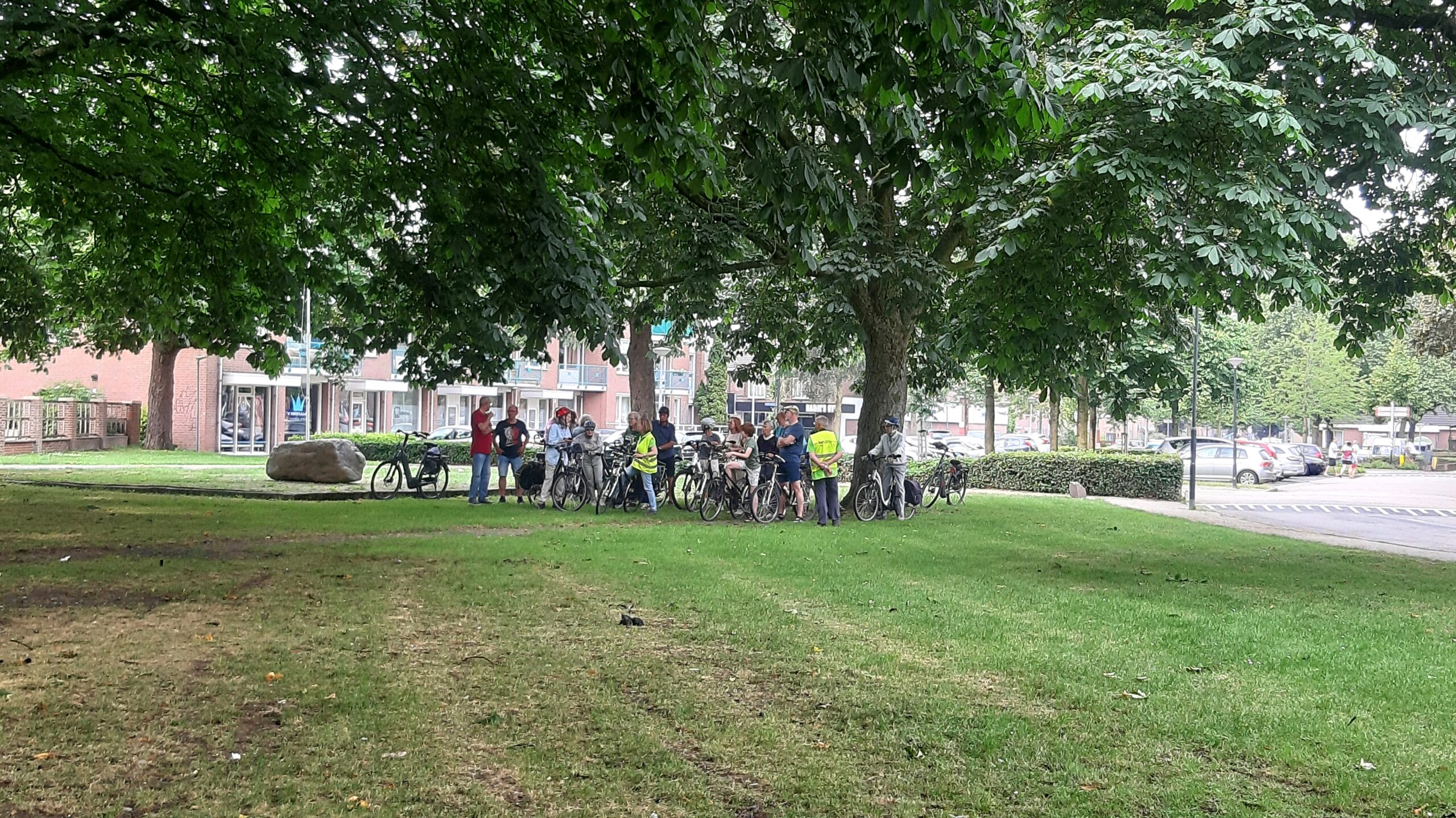 Groep mensen met fietsen onder bomen in een park, naast een parkeerplaats.