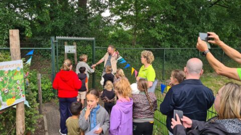 Groep kinderen en volwassenen verzameld bij een afgesloten poort in een bosrijke omgeving.