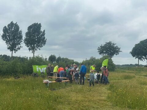 Groep mensen verzameld rond een tafel in een grasveld met groene banners op de achtergrond.