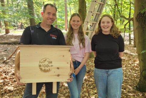 Drie mensen in een bos, één houdt een houten vogelhuis met logo vast.