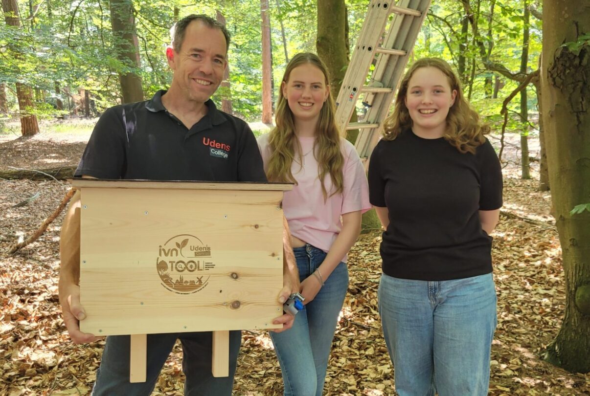 Drie mensen in een bos, één houdt een houten vogelhuis met logo vast.