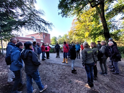 Groep mensen verzameld in een bosrand bij een bakstenen gebouw, luisteren naar een spreker.